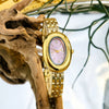 Reloj vintage dorado con esfera rosa y caja dorada junto a una rama seca.
