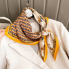 A yellow silk scarf with an orange geometric pattern is tied around a white blazer and displayed on a hanger.