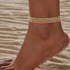 Beaded anklets in pink and green glow-in-the-dark colors worn on a woman's ankle against a sandy background.