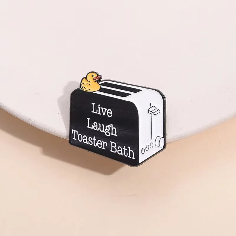 A black and white enamel pin featuring a duck illustration and the text Live Laugh Toaster Bath