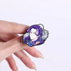 A hand holds a purple-haired anime character brooch lapel pin featuring delicate floral accents.