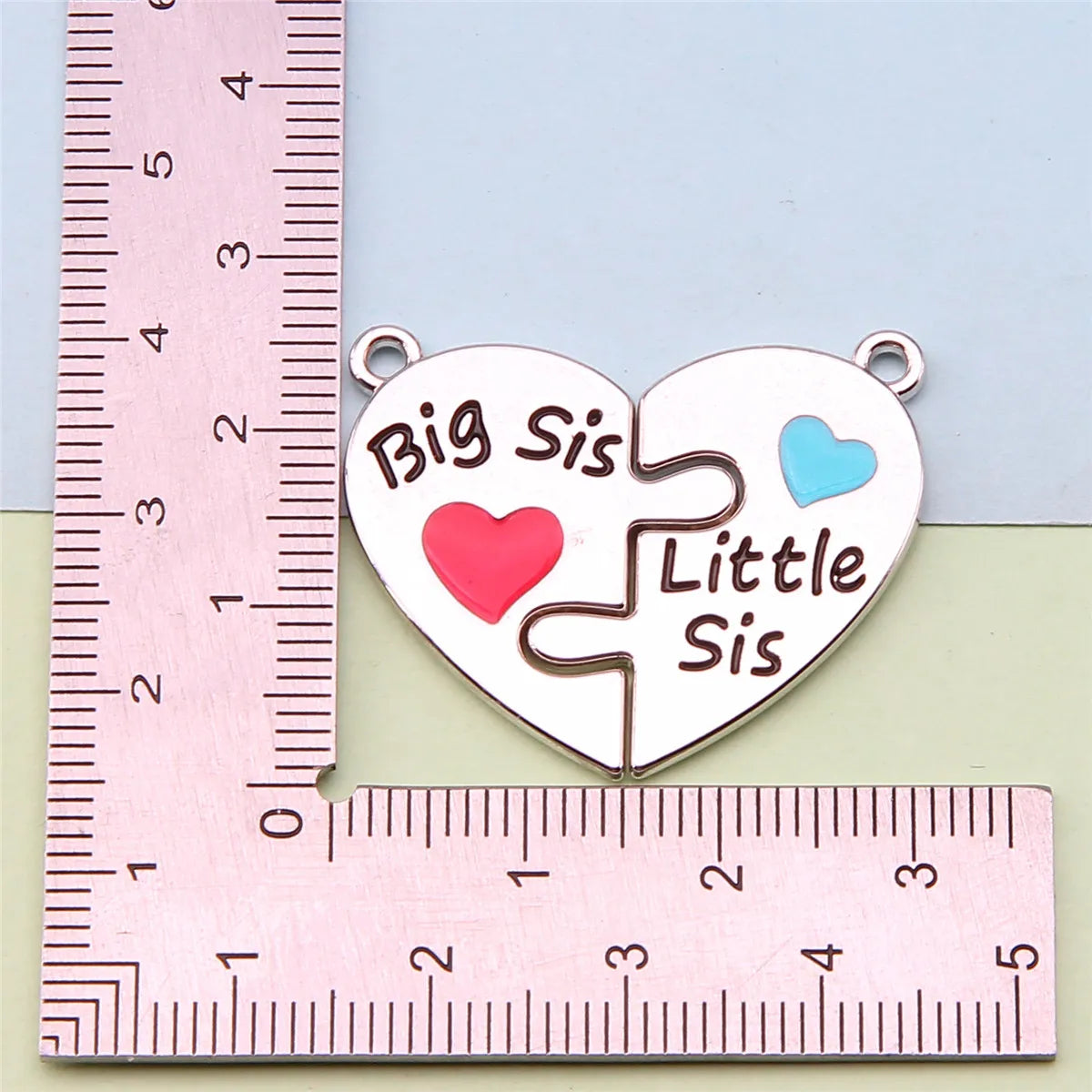 Colgante de collar en forma de corazón con diseño de pieza de rompecabezas y texto 'Big Sis' y 'Little Sis' sobre fondo rosa.