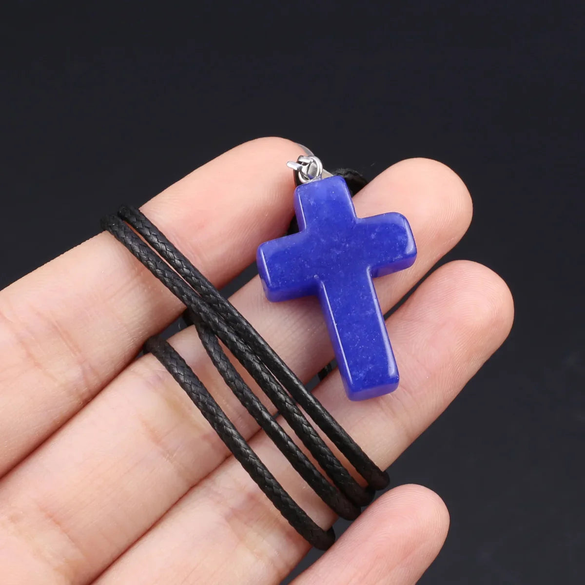 Eine Hand hält eine leuchtend blaue Tigerauge-Agat-Kreuz-Anhänger-Kette vor einem dunklen Hintergrund.
