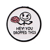 A white enamel pin featuring a simple cartoon face with a pink brain, with the text Hey you dropped this.