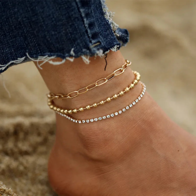 Nahaufnahme einer Hand mit einem goldenen Vintage-Schmetterlings-Herz-Anklet an einem Fuß vor sandigem Hintergrund.