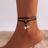 A close-up of a black leather anklet with two silver heart charms worn on a person's ankle.