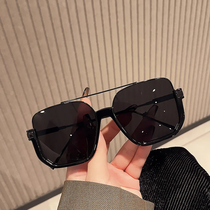 A hand holds a pair of black double bridge oversized square sunglasses against a light background.