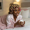 A woman wearing a leopard print hair drying cap is sitting in bed with pink bedding.