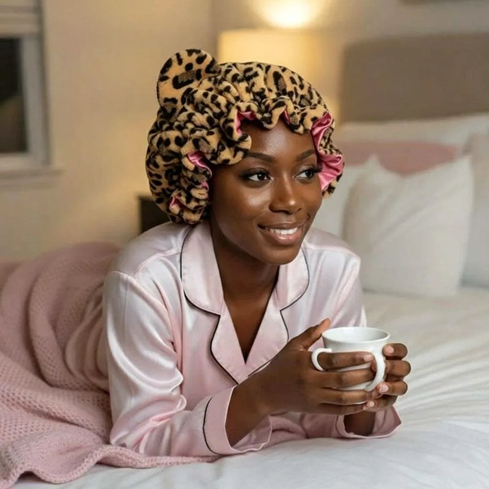 A woman wearing a leopard print hair drying cap is sitting in bed with pink bedding.