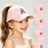 A young girl is smiling while wearing a pink unicorn letter baseball cap and holding several other letters.