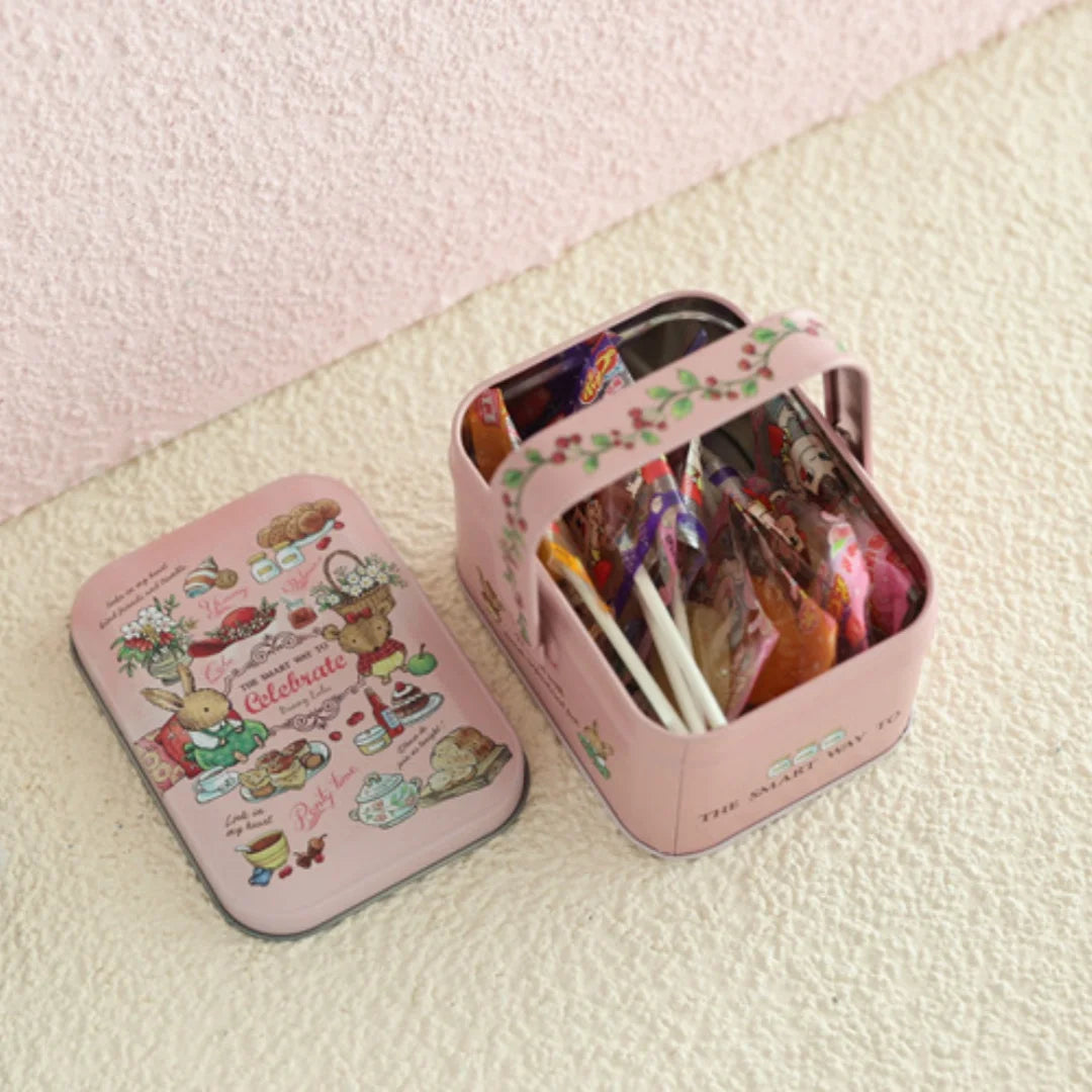 A pink decorative tinplate candy box with a carrying loop is shown resting on a light surface.