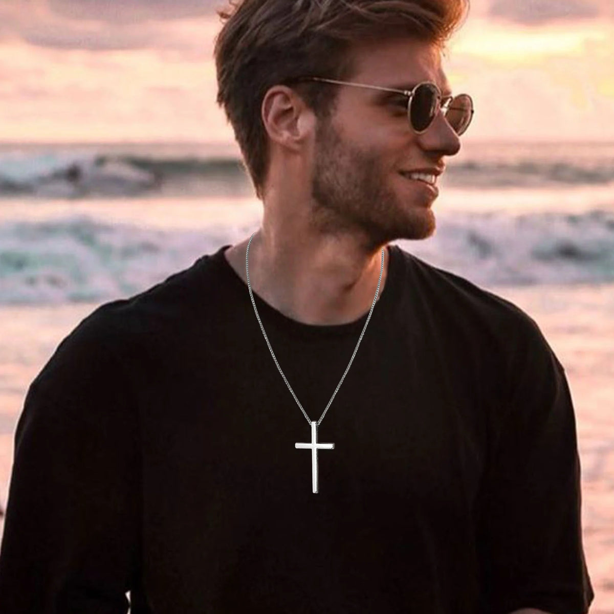 Un hombre con gafas de sol sonríe mirando al mar, llevando un collar de cruz plateado.