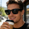 A young man wearing black square sunglasses and holding a coffee cup outdoors.