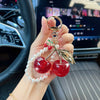 A hand holds a pearl bead key chain decoration featuring red cherries and gold accents against a car interior background.