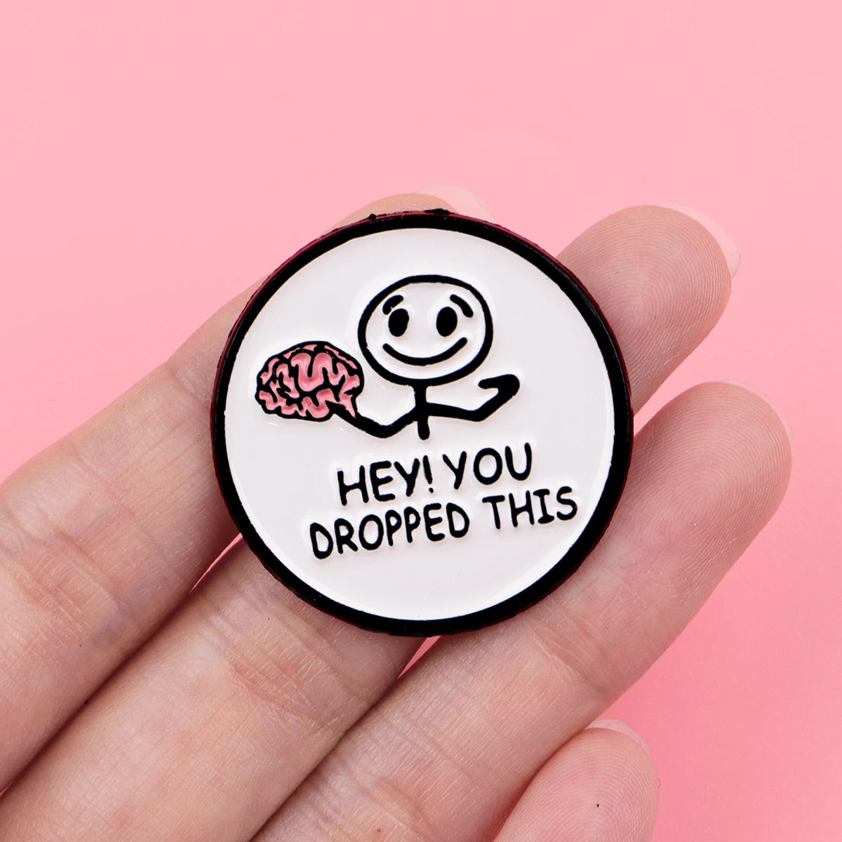A black enamel pin featuring a simple line drawing of a brain and the text Hey you dropped this