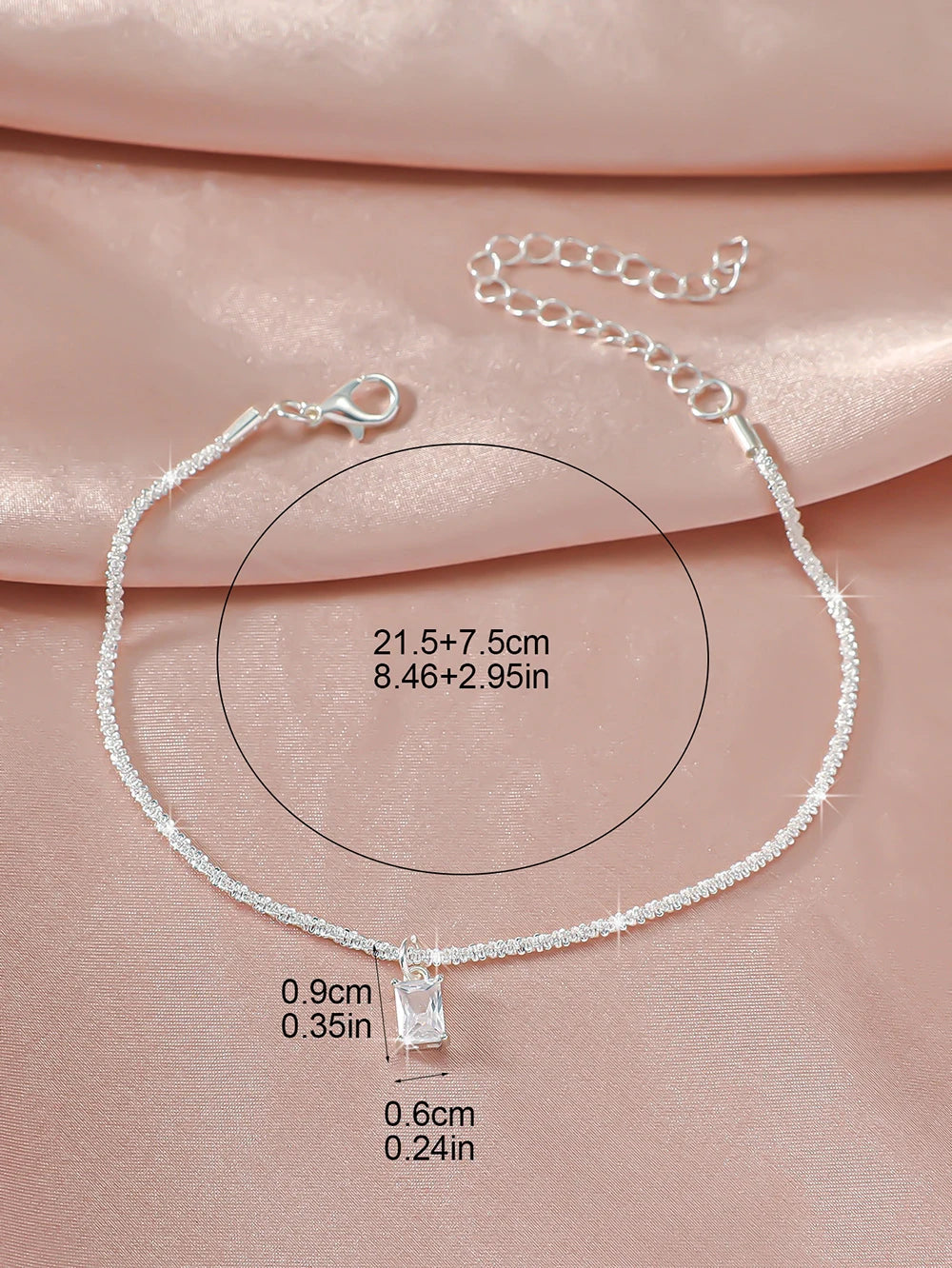 A delicate sparkling zircon heart cross anklet displayed on a pink fabric background.
