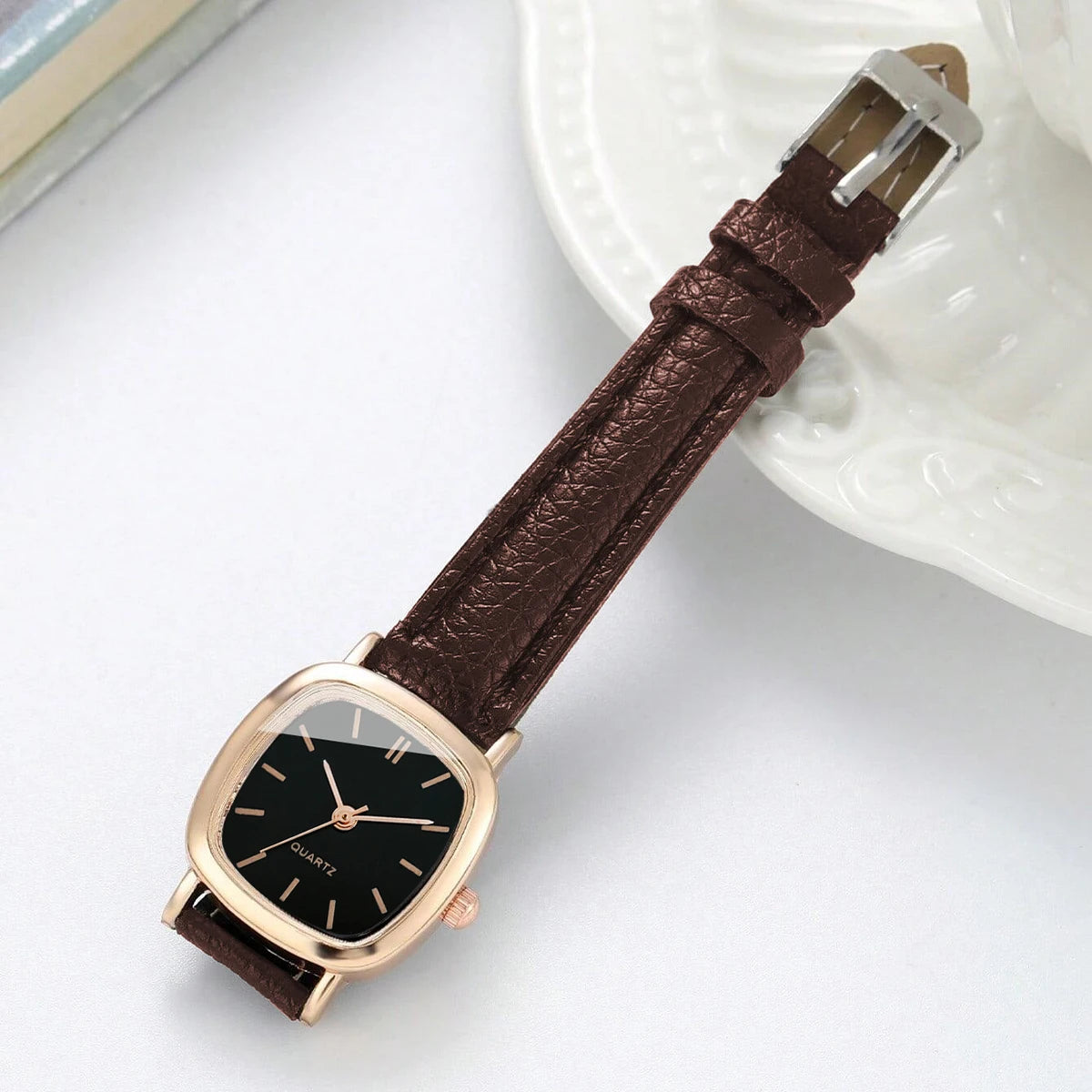 A brown leather watch with a gold-toned square face and a silver clasp resting on a white plate.