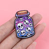 A hand holds a colorful enamel pin featuring purple and blue skulls on a dark background.