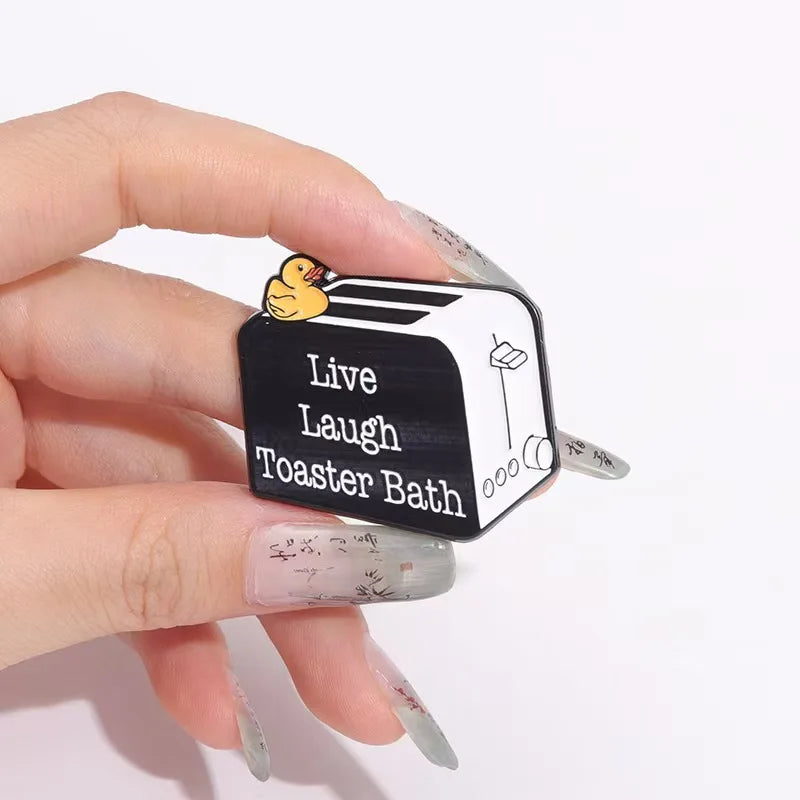 A hand holds a black enamel pin featuring a duck and the text Live Laugh Toaster Bath