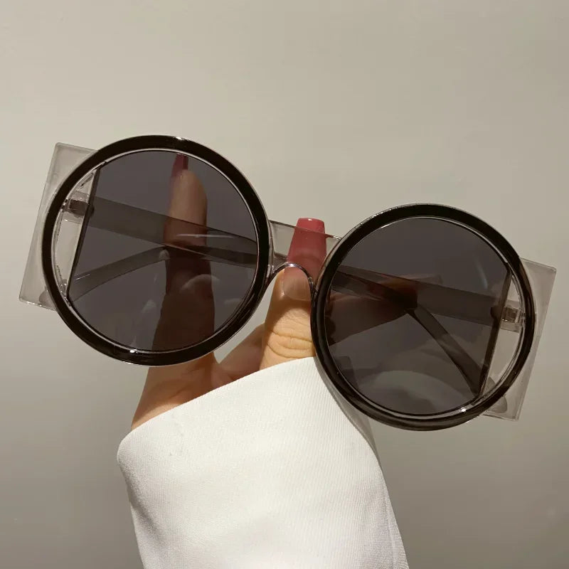 A hand holds a pair of round double color fashion sunglasses with dark lenses and clear frames.