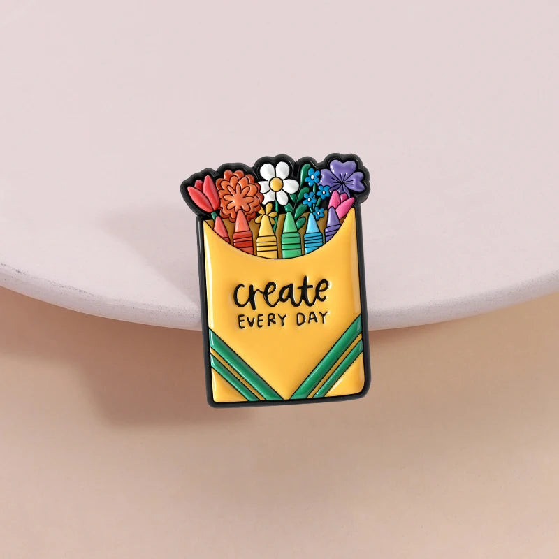 A yellow enamel brooch featuring a colorful arrangement of flowers and the text create every day.
