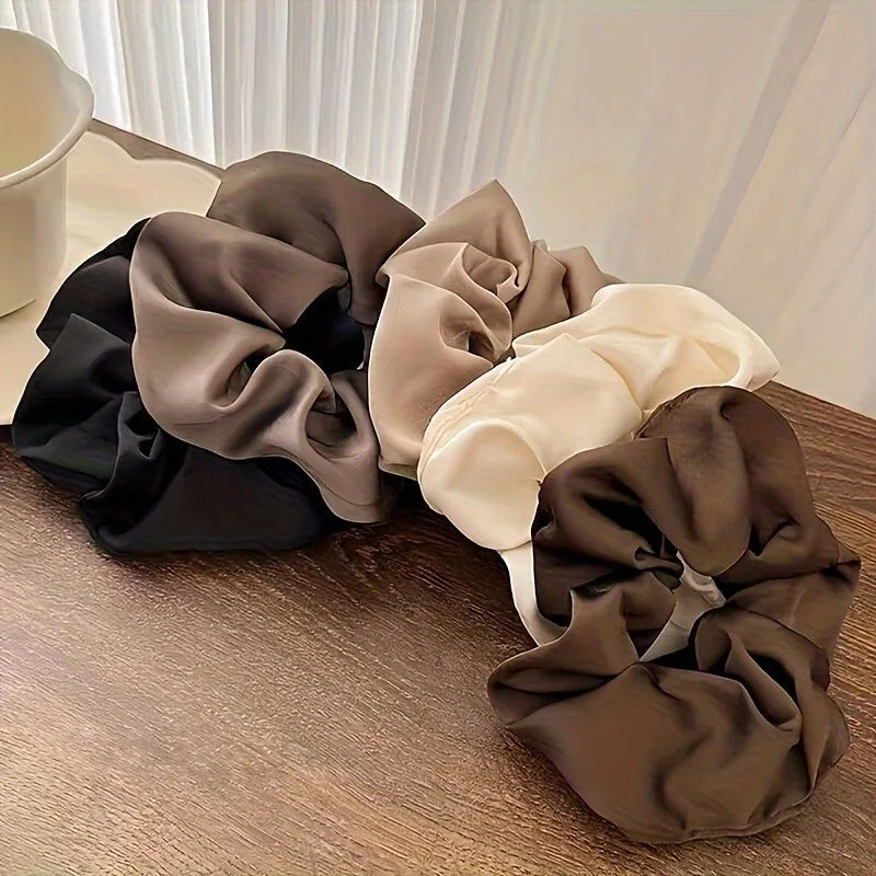 A collection of five elegant silk scrunchie hair ties in shades of black, gray, beige, and brown are displayed on a wooden su