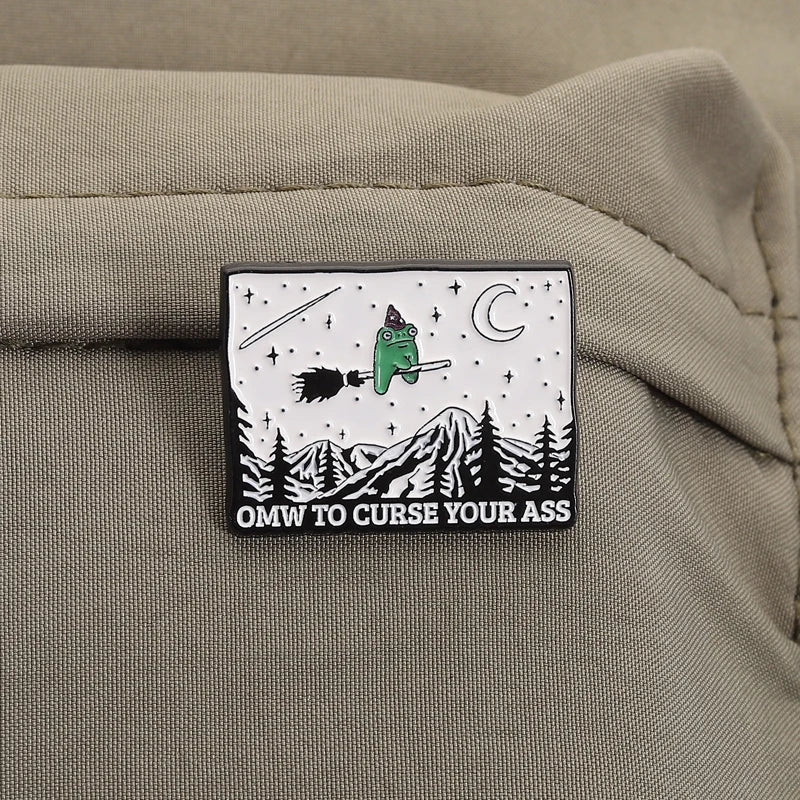 An enamel pin featuring a green frog wizard with a broom and a crescent moon over a snowy mountain landscape.