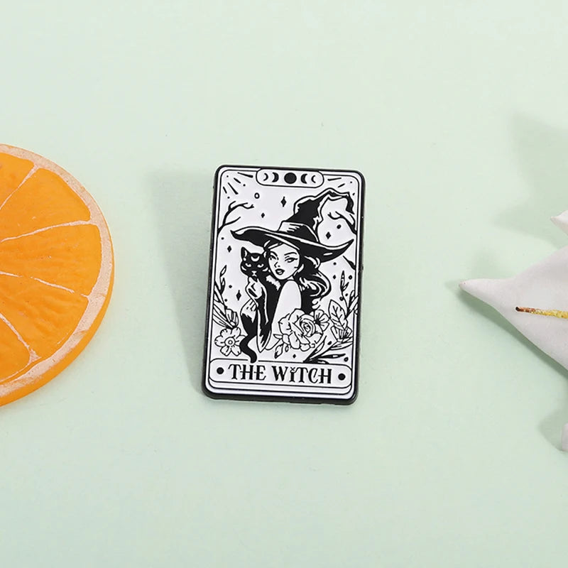A black tarot card enamel pin featuring a witch with a pointed hat and flowers, displayed on a light green surface.