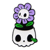 An enamel pin featuring a purple flower surrounding a black skull design.
