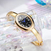 A gold-toned quartz watch with a black face and blue hands is displayed on a clear, iridescent glass surface.