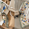 A white silk scarf with a floral print featuring blue and yellow flowers is displayed on a beige fabric and next to a leather