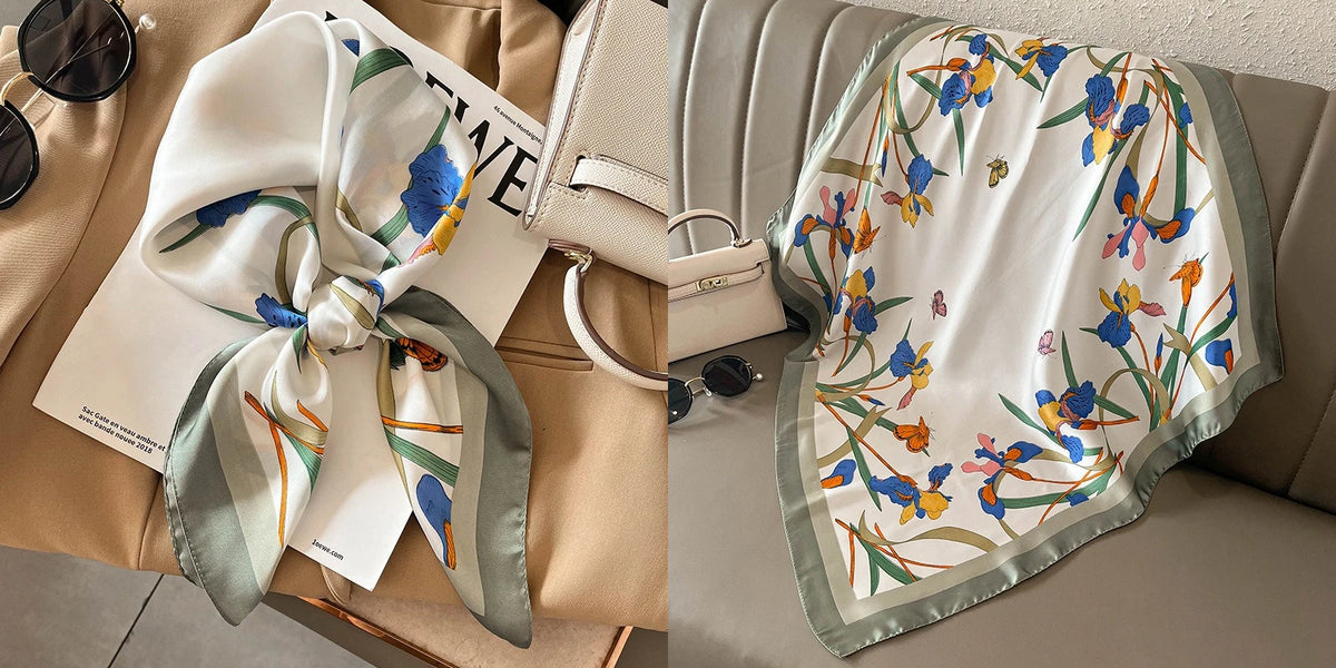 A white silk scarf with a floral print featuring blue and yellow flowers is displayed on a beige fabric and next to a leather