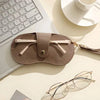 A beige PU leather eyewear storage case resting on a white surface next to a laptop keyboard and eyeglasses.