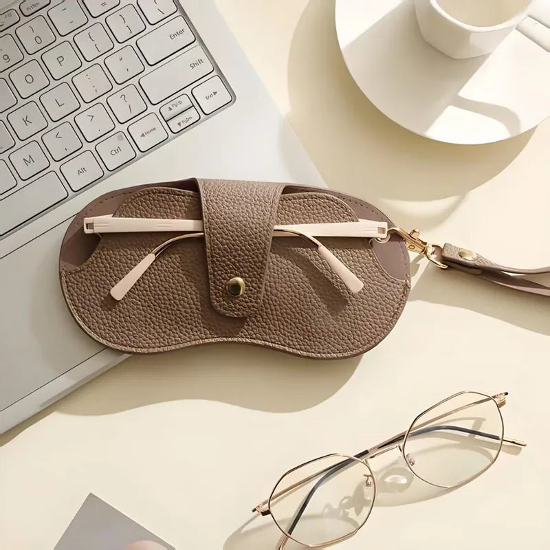 A beige PU leather eyewear storage case resting on a white surface next to a laptop keyboard and eyeglasses.