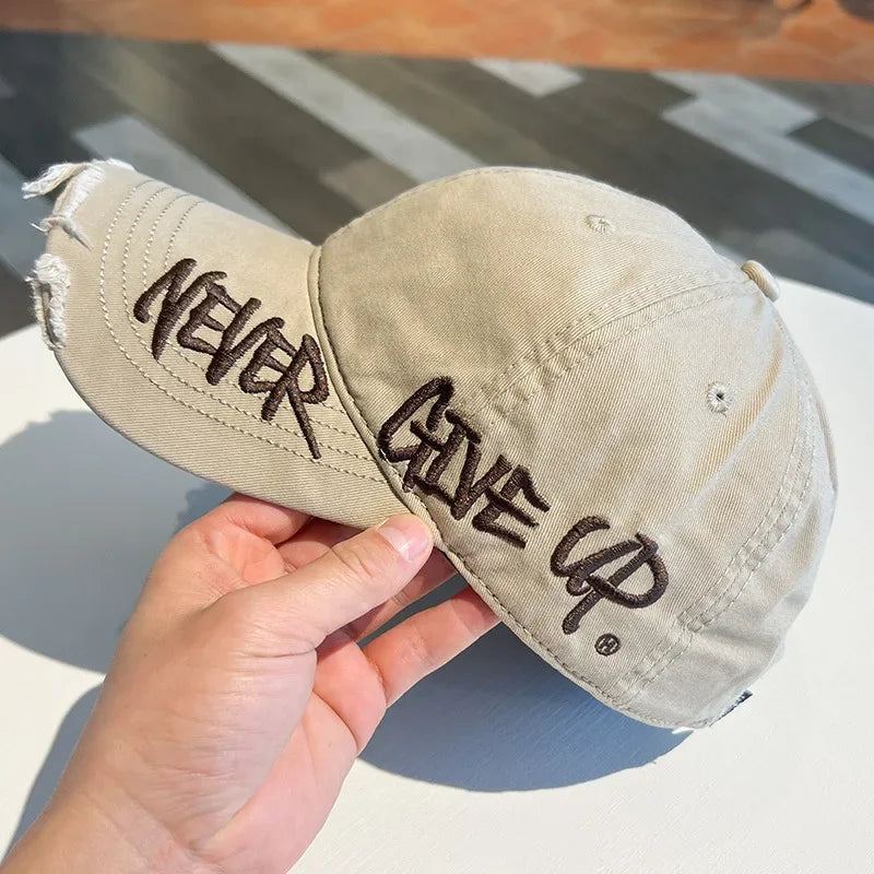 A hand holds a beige vintage washed cotton baseball cap with embroidered text on the front.