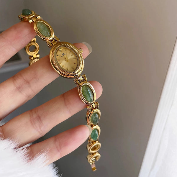 A hand holds an elegant gold-toned wristwatch featuring green jade quartz stones on a gold band.