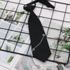 A black pre-tied chain necktie is displayed on a grid background of greenery and paper.
