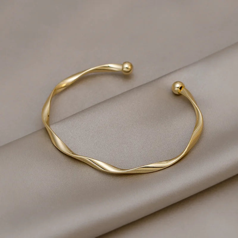 A minimalist gold-tone twisted cuff bangle resting on a neutral gray fabric.