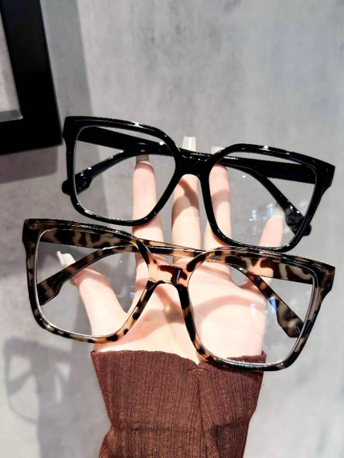 A hand holds a pair of black-framed reading glasses with tortoiseshell patterns against a light gray background.