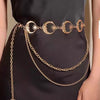 Eine Frau in einem schwarzen Kleid mit goldener Boho-Mond-Fransen-Taillenkette und übereinanderliegenden goldenen Ketten.