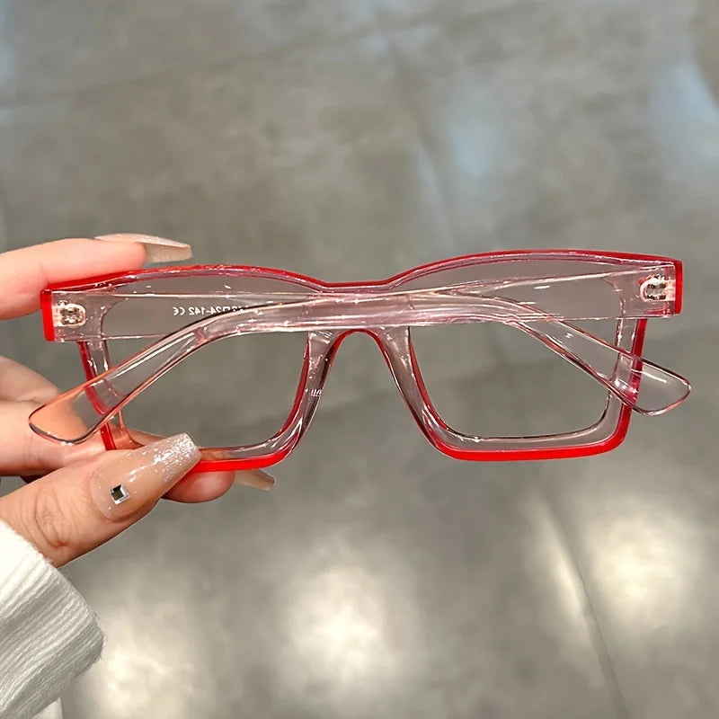 A hand holds a pair of red plastic costume glasses with square frames against a blurred gray background.
