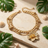 A gold chain anklet featuring a floral pendant rests on a light surface surrounded by green tropical leaves and seashells.