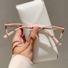 Una mano sostiene unas gafas de computadora con marco rosa y estilo ojo de gato metálico contra un cuaderno de cuero blanco.