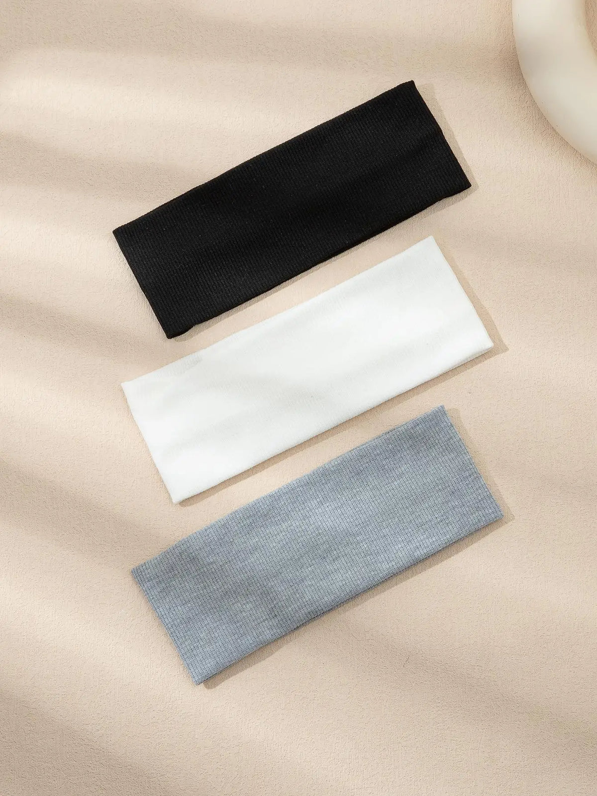 A pack of three solid color fashion headbands in black, white, and gray laid diagonally on a light beige background.