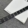 A black PU leather fashion belt with silver grommets is laid across a white card on a gray textured surface.