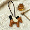 A leather horse pendant charm with a gold-colored wing is displayed on a cream knitted fabric next to honey and a wooden dipp