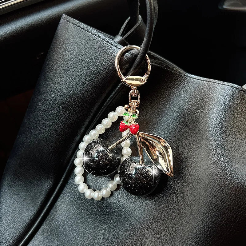 A gold-toned key chain decoration featuring a red cherry and black apples adorned with white pearls.