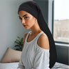 A woman wearing a black jumbo dreadlock cap and a white top is shown indoors.