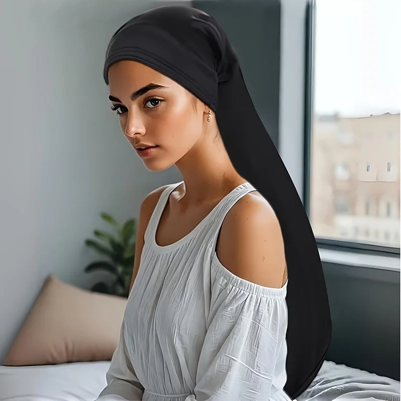 A woman wearing a black jumbo dreadlock cap and a white top is shown indoors.