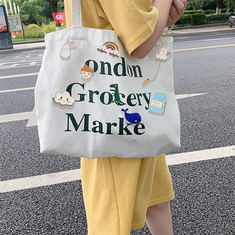 A white tote bag featuring green text that reads Ondon Grocery Marke with various cute animal and plant metal pin badges